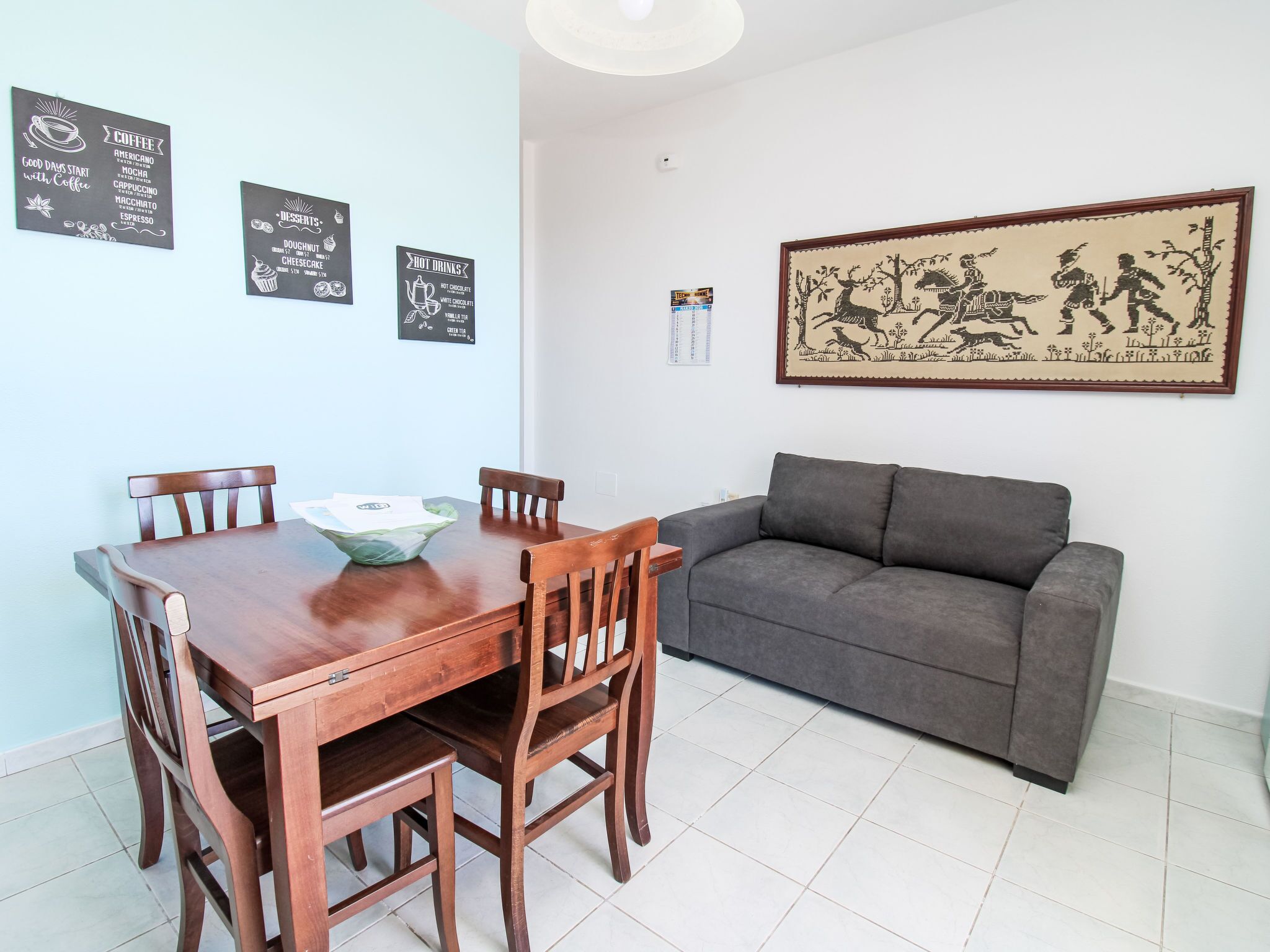 A dining area with a wooden table and chairs, a grey sofa, and wall decorations including a framed picture and coffee-related signs.