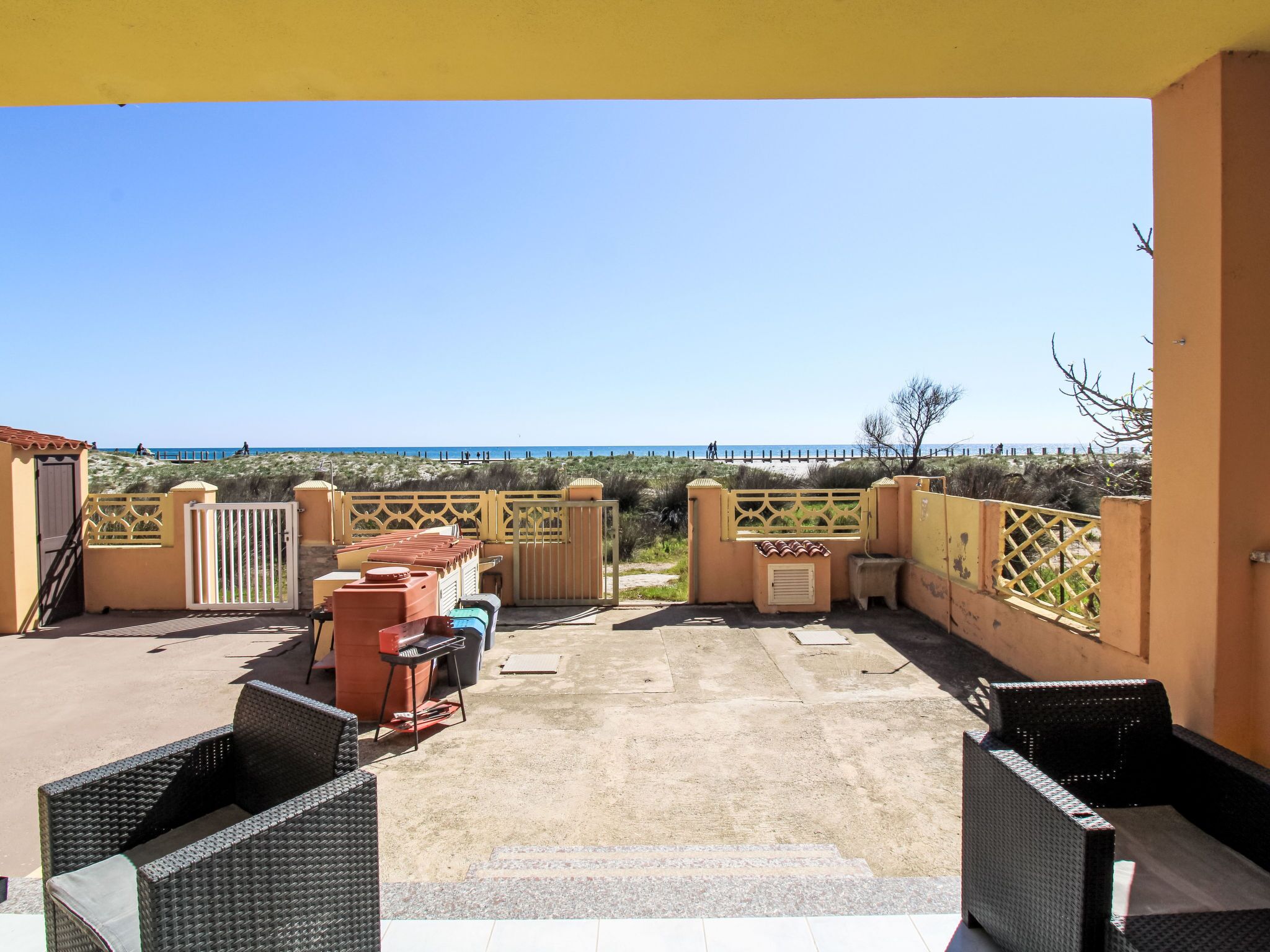A covered patio with wicker furniture, a view of the beach, and a gate leading to the beach.
