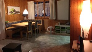 A cozy room with wooden walls and a wooden ceiling. There is a dining table with chairs, a small stool, and a lamp. A window with lace curtains is visible, and there are storage bins against the wall.