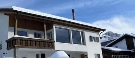 Ein zweistöckiges Gebäude mit Balkon, großen Fenstern und einem mit Schnee bedeckten Dach.