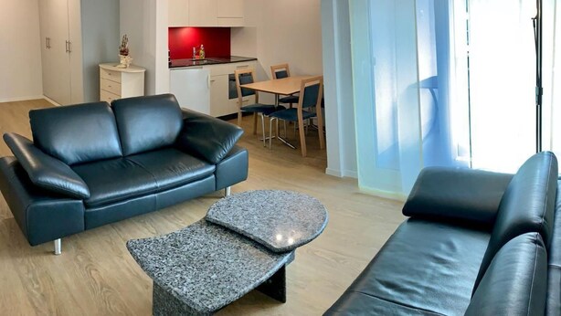 Ein modernes Wohnzimmer mit einer schwarzen Ledersofa, einem kleinen Stehtisch und einem Essbereich mit Holztisch und Stühlen.