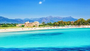 Paesaggio costiero con mare cristallino, spiaggia sabbiosa e un edificio storico sullo sfondo montuoso.