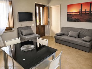Een woonkamer met een zwarte eettafel, twee grijze banken, een aan de muur bevestigde televisie en een schilderij van een zonsondergang.