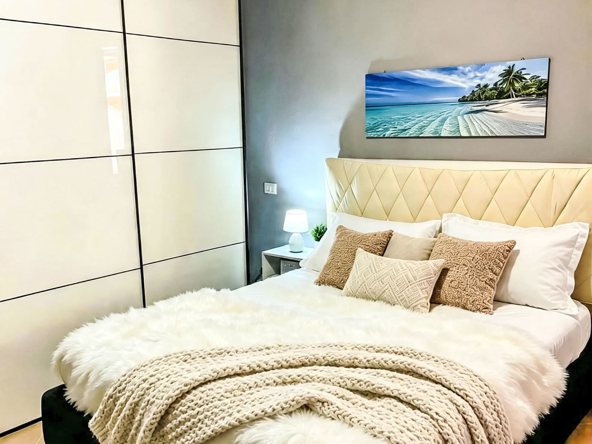 A bedroom with a bed, pillows, a nightstand, a lamp, and a painting of a beach scene.