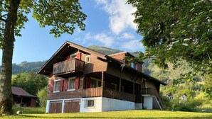 A two-story wooden house with a balcony, surrounded by a grassy yard and trees, set against a mountainous backdrop. - Chalet Bacchus (EG) by Interhome (Sörenberg)