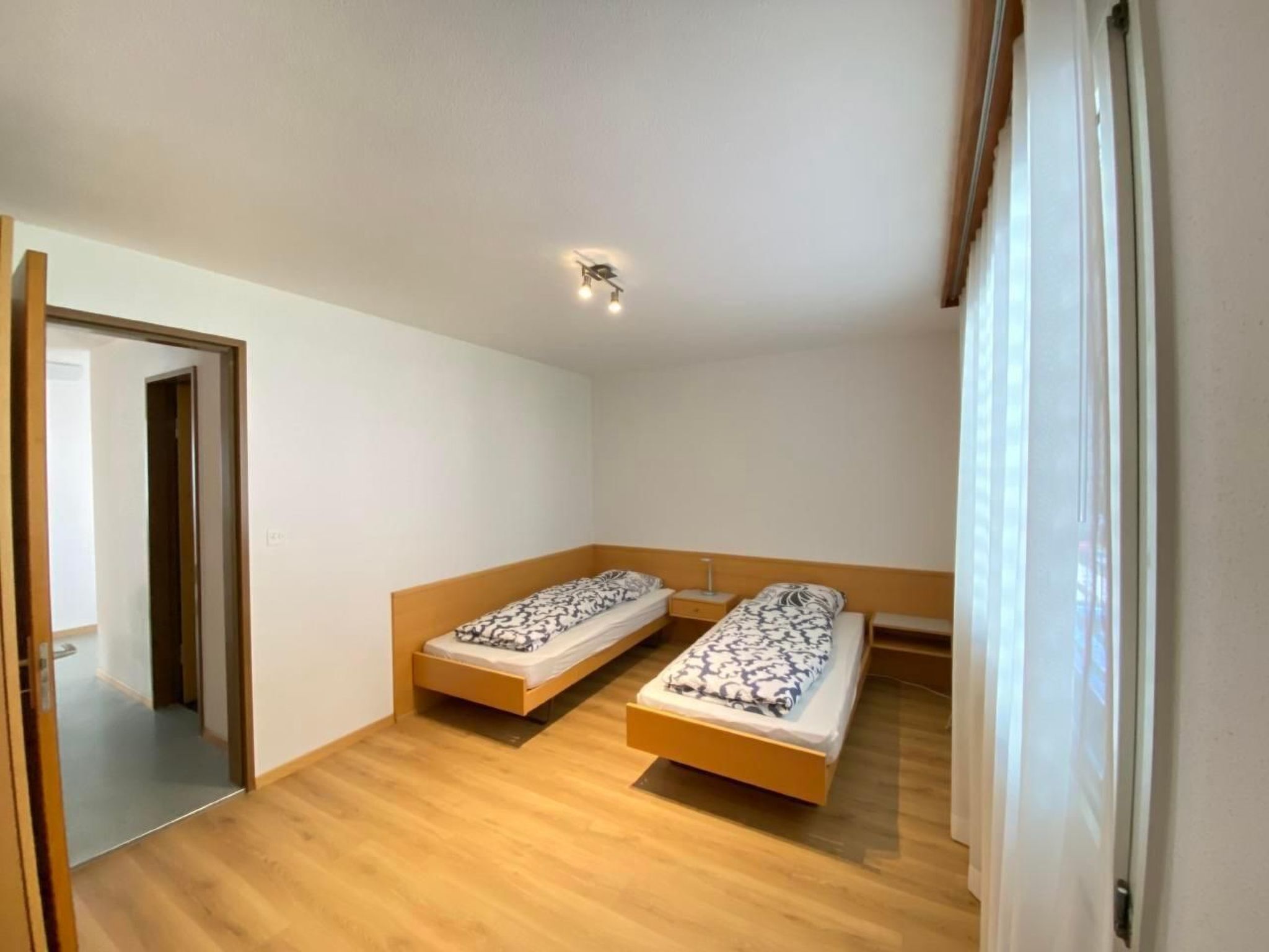 A small, bright room with two single beds, wooden flooring, a ceiling light, and a door leading to another room.
