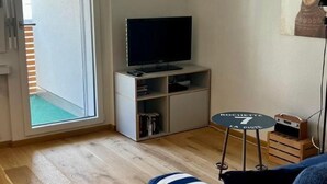 A living room with a television on a stand, a small round table, a sofa, and a door leading to a balcony.