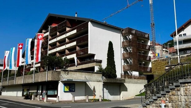 A modern building with multiple balconies, a construction crane in the background, and flags on the left.