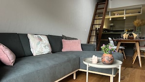 Ein modernes Wohnzimmer mit einem grauen Sofa, einem kleinen runden Tisch mit einer Vase und einem Keramikvogel, sowie einem Holzessttisch mit Stühlen.