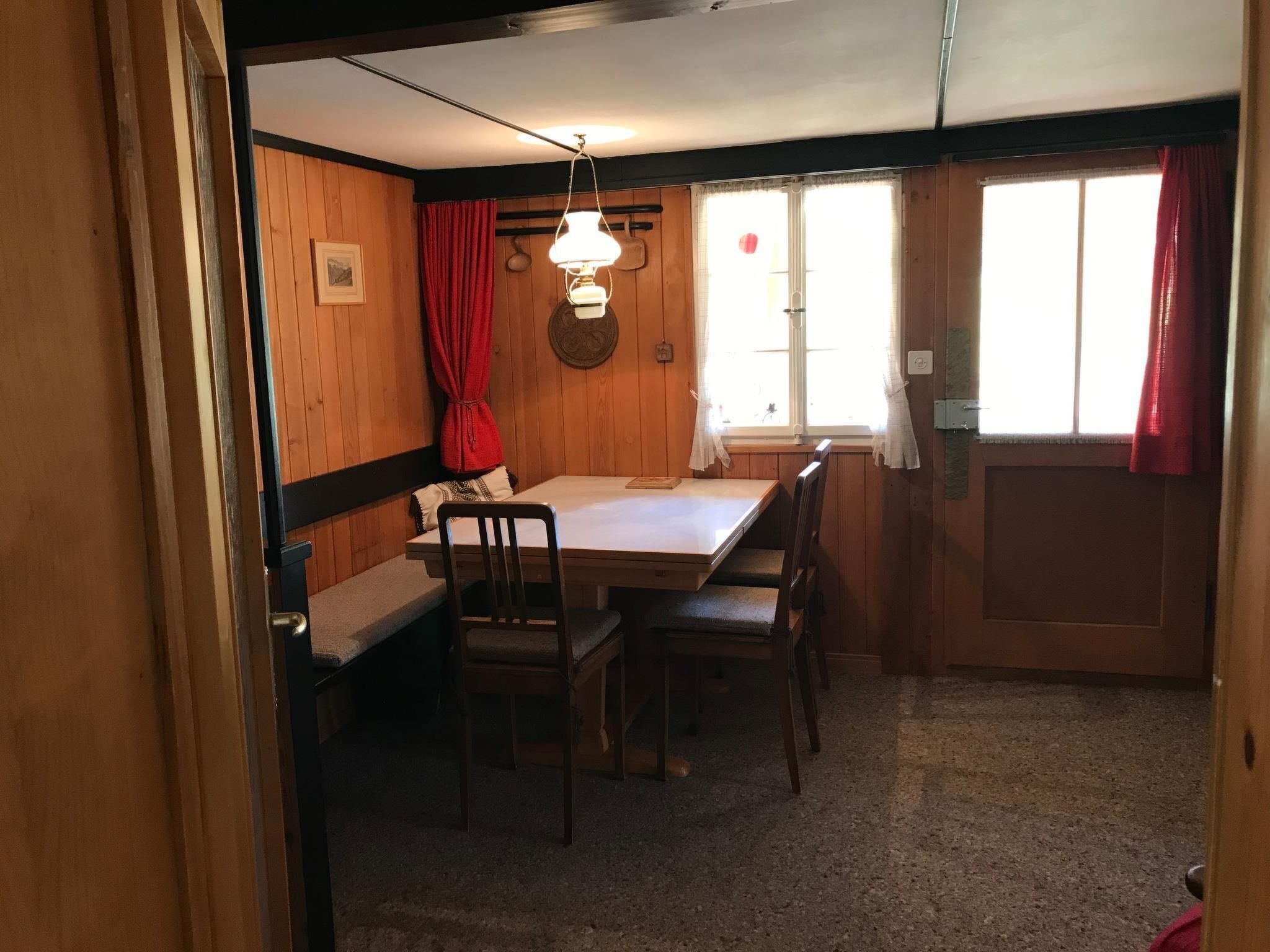 A small, cozy room with wooden walls and a single window with red curtains. There is a table with chairs and a hanging light fixture.