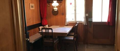 A small, cozy room with wooden walls and a single window with red curtains. There is a table with chairs and a hanging light fixture.