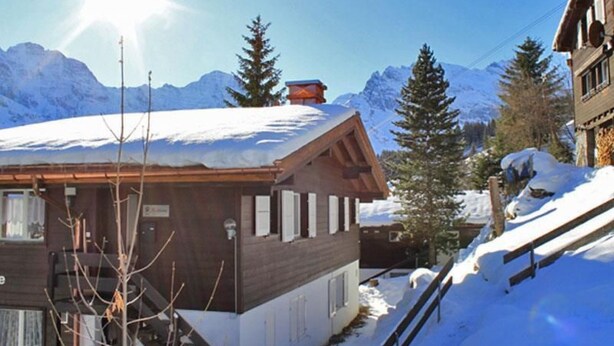 Una cabaña de madera con techo inclinado, rodeada de montañas y árboles cubiertos de nieve.