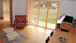 Un salón con un sillón de cuero rojo, una mesa de comedor de madera con un tazón y vasos, un televisor y un balcón con persianas de madera.