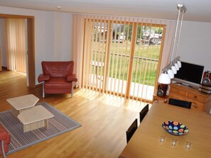 Ein Wohnzimmer mit einem roten Ledersessel, einem Holzesstisch mit Schüssel und Gläsern, einem Fernseher und einem Balkon mit Holzhörchen.