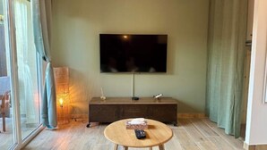 Ein Wohnzimmer mit einem großen Flachbildfernseher an der Wand, einem Holztisch, einer dunkelbraunen Holzkommode und einer Glastür mit Vorhängen.