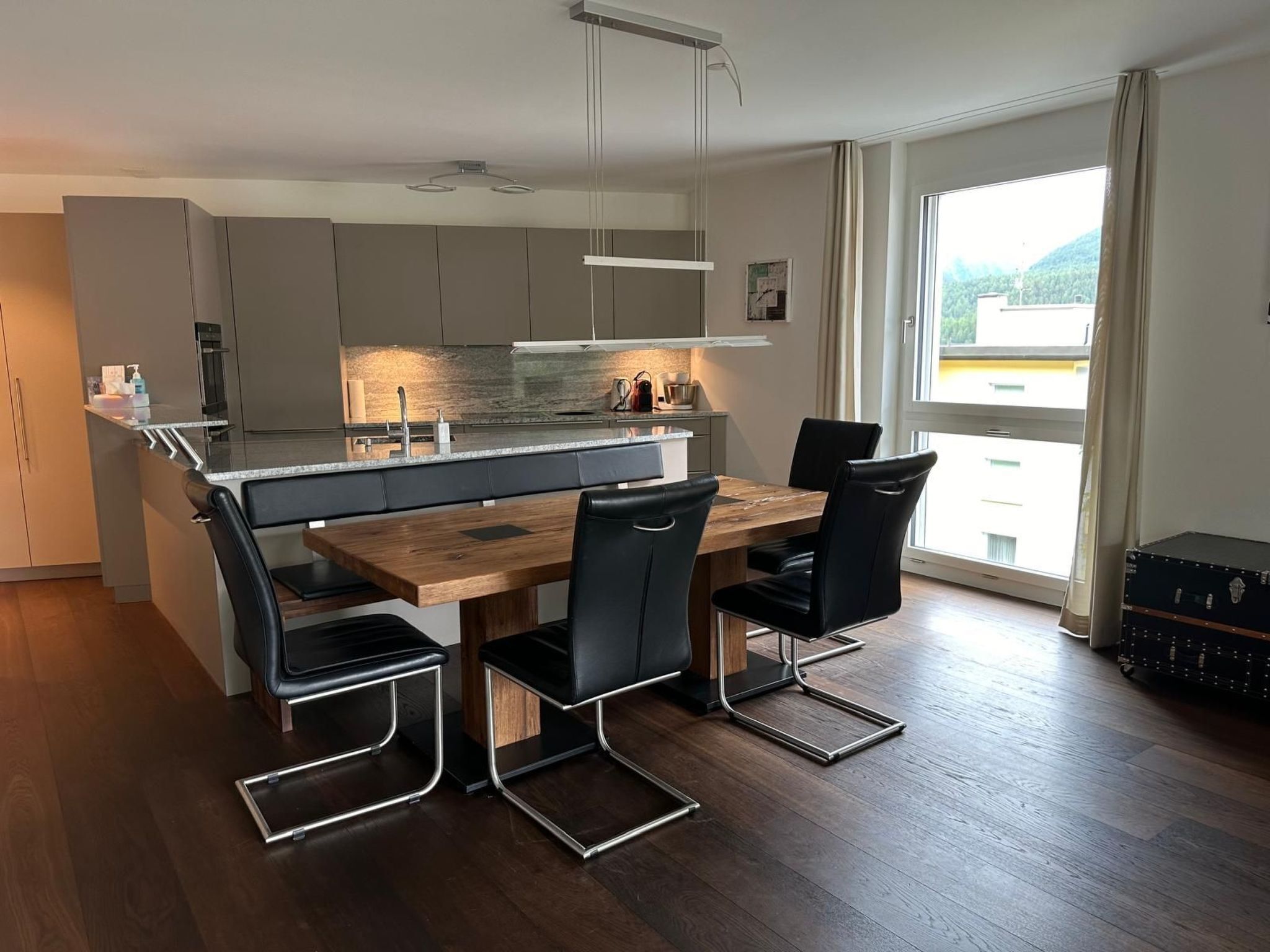 A modern kitchen with a central island, a dining area with a wooden table and black chairs, and a large window offering a view of the outdoors.