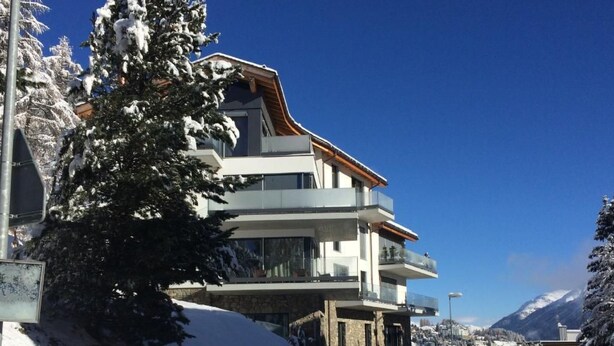 Ein modernes, mehrstöckiges Gebäude mit Balkonen, umgeben von schneebedeckten Bäumen und Bergen im Hintergrund.
