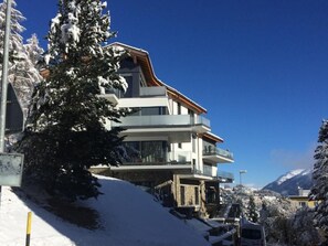 En modern, flervåningsbyggnad med balkonger, omgiven av snötäckta träd och berg i bakgrunden.