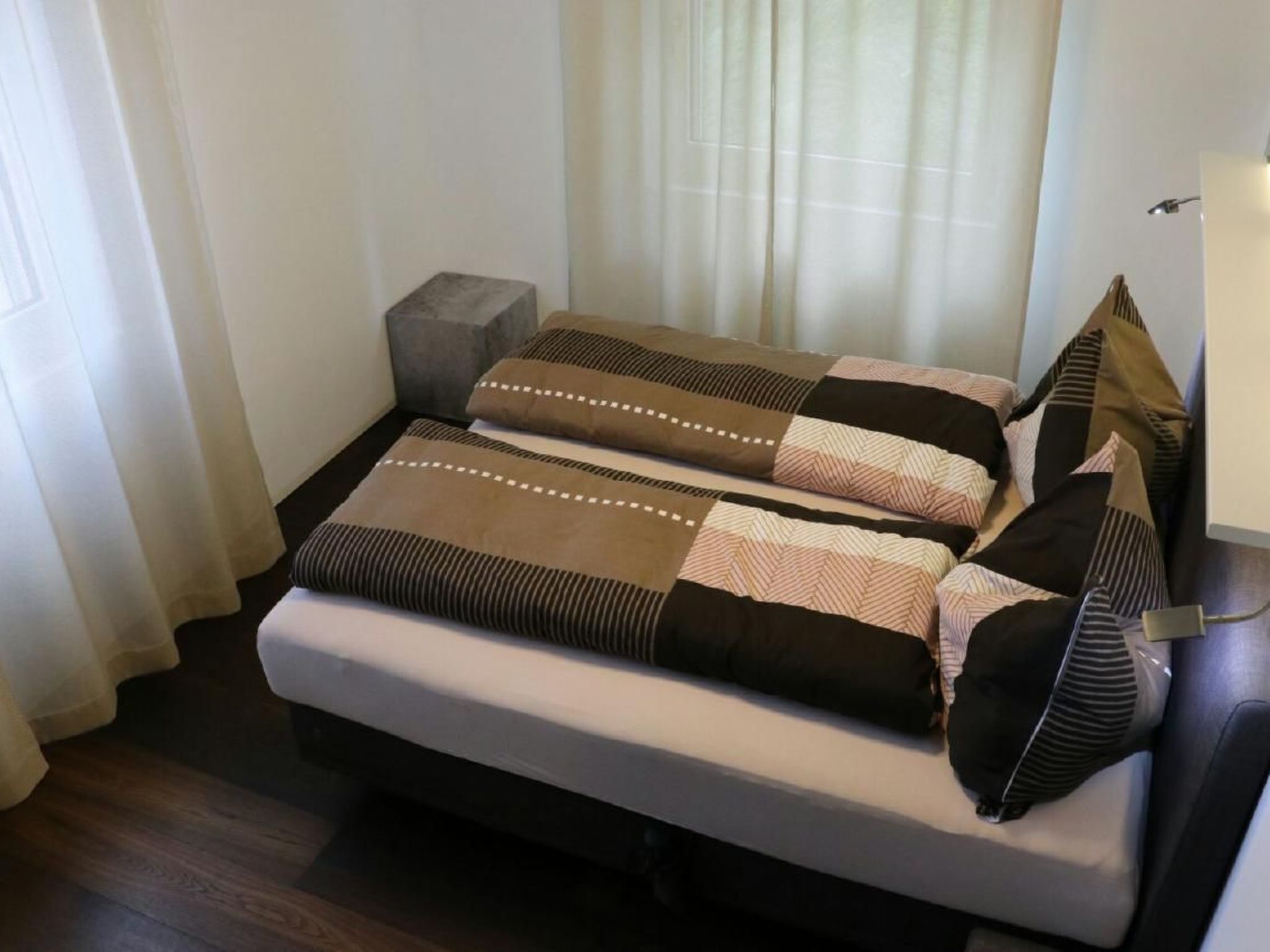 A bed with a white mattress and brown striped bedding, a small gray nightstand, and a window with sheer curtains.