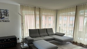Ein modernes Wohnzimmer mit einem Ecksofa, einer Stehlampe und einem großen Fenster mit durchsichtigen Vorhängen.