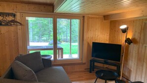 L’intérieur d’un chalet accueillant, avec un canapé, un téléviseur posé sur un meuble, une petite table et une grande fenêtre donnant sur un espace extérieur verdoyant.