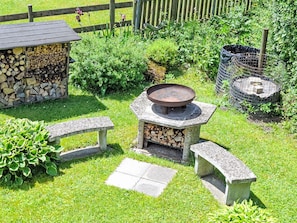 Un área de cocina al aire libre de piedra con un hogar central, rodeada por una valla de madera, un banco de piedra y un cobertizo pequeño con leña apilada.
