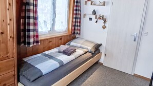 Yhden hengen sänky päätyineen, ikkuna verhoineen, ovi ja seinälle kiinnitetyt hyllyt, joilla on koriste-esineitä.