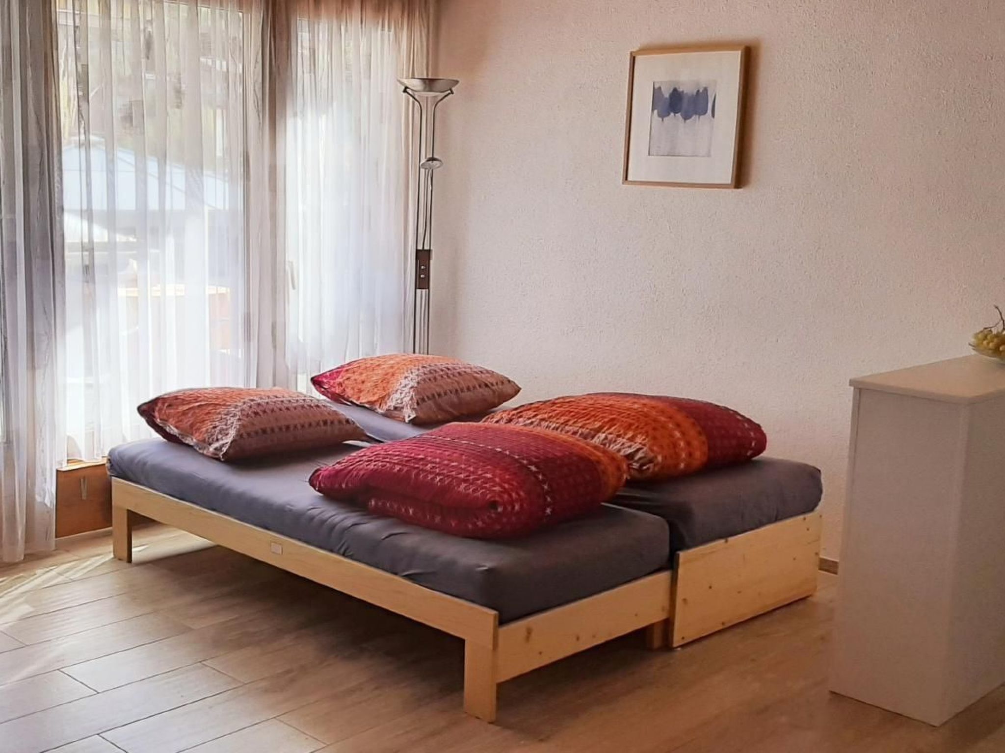 Een enkel bed met houten frame en grijs matras, met meerdere kleurrijke kussens. Een vloerlamp staat naast het bed, en aan de muur hangt een ingelijst beeld.