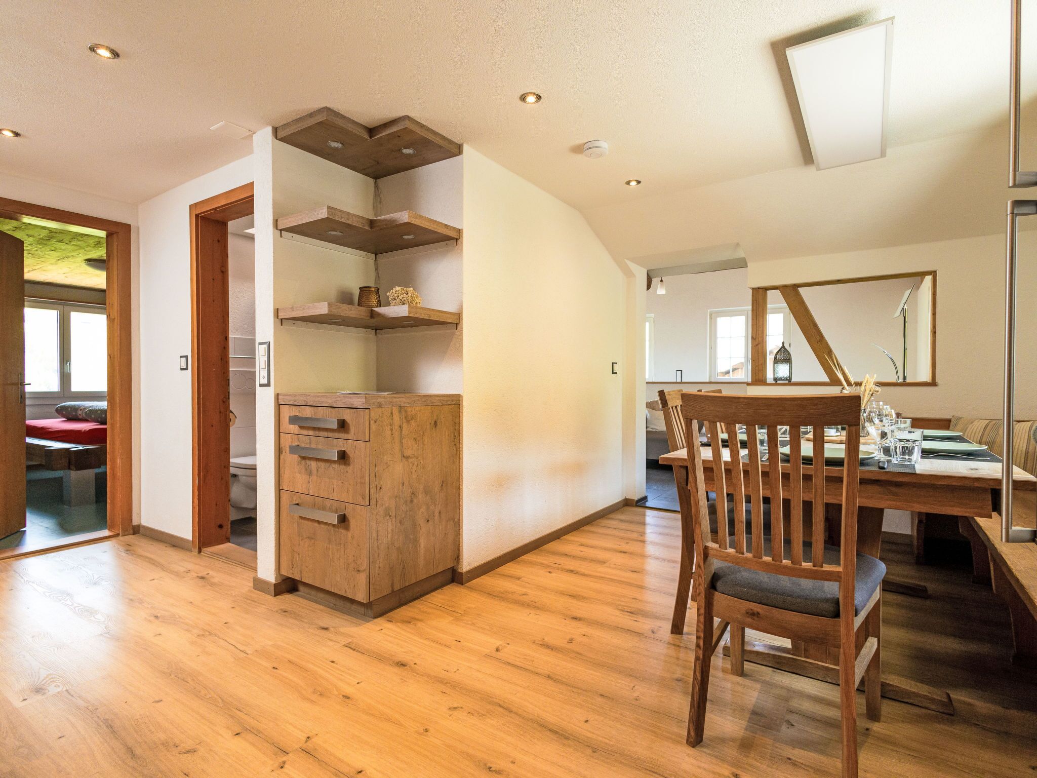 A spacious room with wooden flooring, a wooden cabinet with drawers, a dining table with chairs, and a large mirror on the wall.