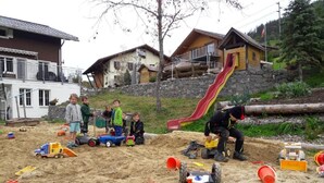 A sandy play area with children and adults, surrounded by houses with balconies and a red slide. - Schrattewohnung Birkenhof (4-Bett) by Interhome (Sörenberg)
