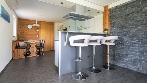 A modern kitchen with a central island, bar stools, a wooden dining area, and a stone accent wall. - Schrattewohnung Birkenhof (4-Bett) by Interhome (Sörenberg)