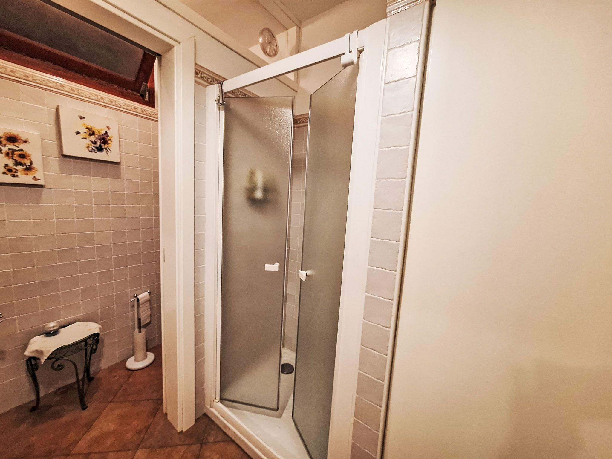 A bathroom with a glass-enclosed shower, white tiled walls, a small table with a towel, and two framed pictures on the wall.