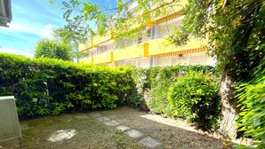 A courtyard with a paved path, dense greenery, and a building in the background. - VILLA CASTORINI - B2 4+0 - B3003C (Bibione Pineda)
