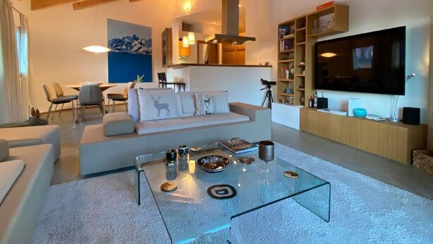 A modern living room with a glass coffee table, a sofa with decorative pillows, a dining area with chairs, a mounted TV, and a bookshelf. - Chesa Baia by Interhome (La Punt-Chamues-ch)