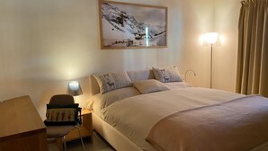A bedroom with a large bed, a nightstand with a lamp, a chair, and a framed picture of a snowy mountain scene. - Chesa Baia by Interhome (La Punt-Chamues-ch)