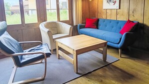 Une pièce dotée d’un plancher en bois, d’un canapé bleu agrémenté de coussins rouges, d’un fauteuil blanc, d’une table basse en bois et de grandes fenêtres offrant une vue sur la verdure et les bâtiments.