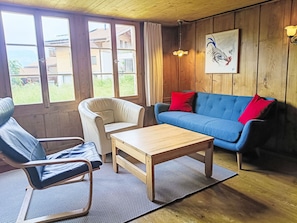 Una habitación con piso de madera, un sofá azul con cojines rojos, un sillón blanco, una mesa de centro de madera y ventanas amplias con vista a zonas verdes y edificios.