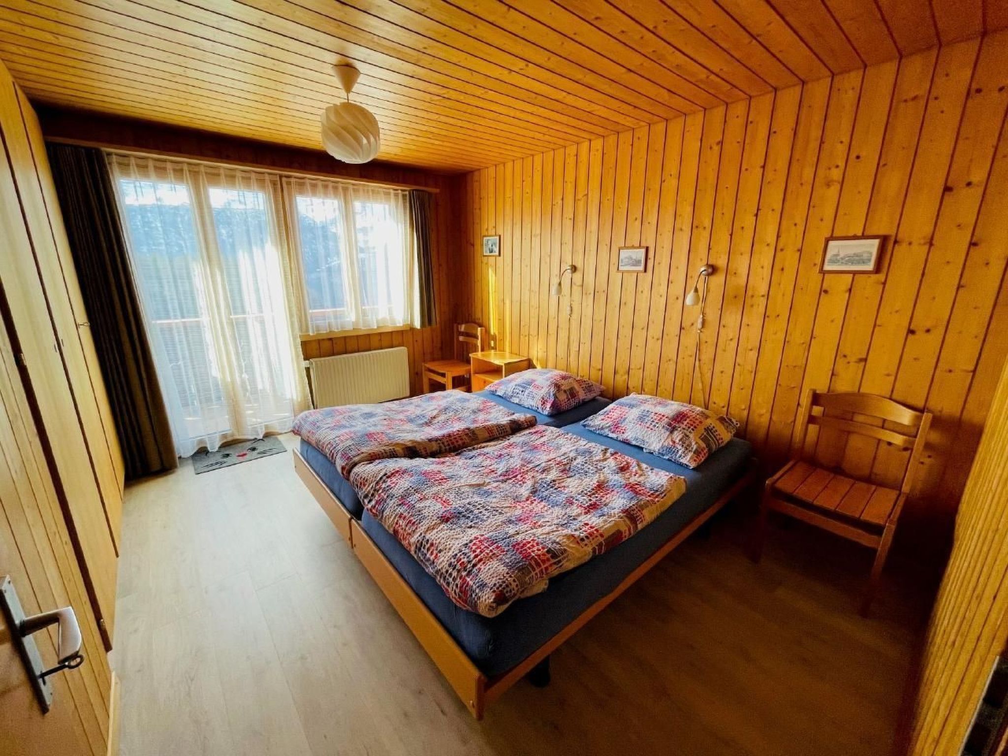 A cozy wooden cabin bedroom with two beds, a window with curtains, a wooden chair, and a small table.