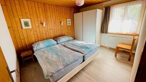 Makuuhuoneessa on puinen katto, sinivalkoinen peitto sängyllä, puinen tuoli ja ikkuna, jossa on verhot.