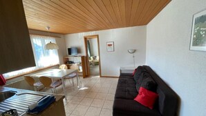 Una sala de estar con un sofá negro, una mesa de comedor con sillas, un televisor y una ventana con cortinas.