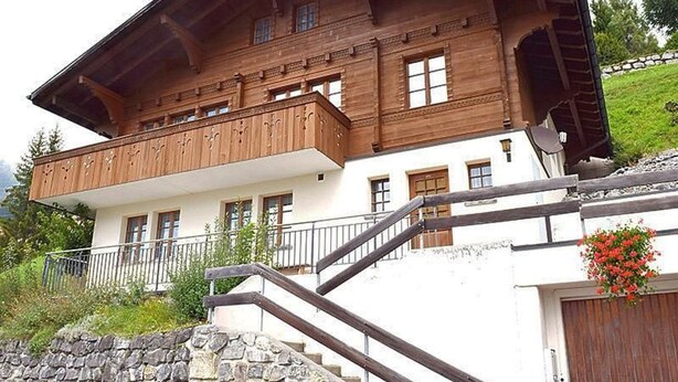 Un chalet de dos pisos con balcón de madera, escalones de piedra y un muro de piedra.