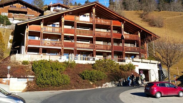 Un edificio de varios pisos con balcones, un telesilla al fondo y un estacionamiento con coches.