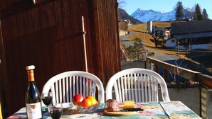 Una mesa con una botella de vino, una copa, un bol de fruta y una tabla de quesos en un balcón con vistas a un paisaje montañoso.