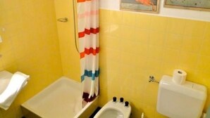 Kylpyhuone, jonka seinät ovat keltaiset, ja jossa on suihkuverho, wc-istuin, bidee sekä kaksi kehystettyä kuvaa seinällä.