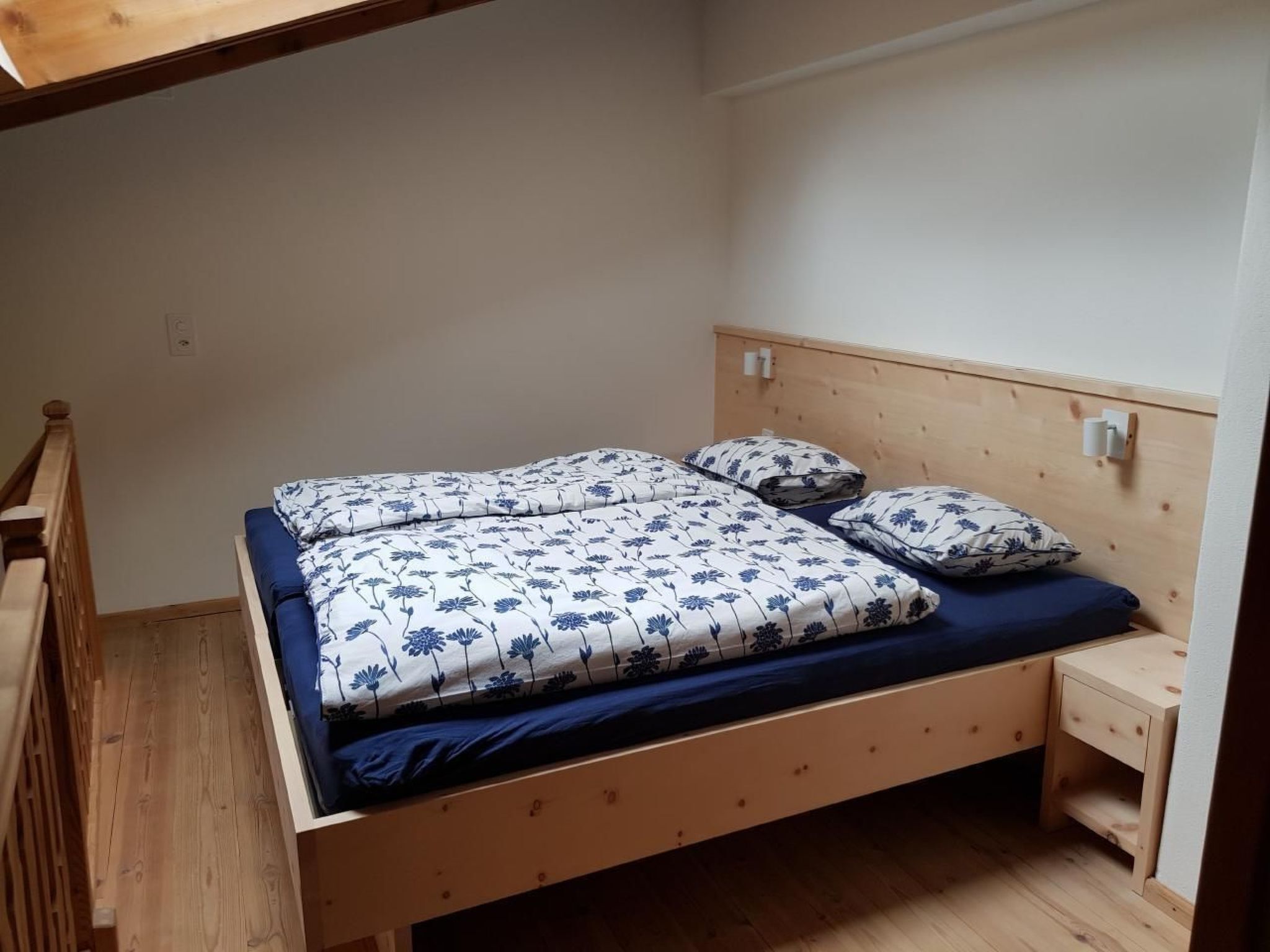 Uma cama de solteiro com estrutura e cabeceira de madeira, um criado-mudo pequeno e uma mesa de cabeceira com abajur.