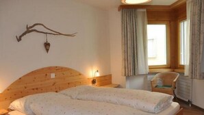 A cozy bedroom with a wooden headboard, two beds with white linens, a nightstand, a wall-mounted heart decoration, a window with curtains, and a wicker chair. - Apartment  by Interhome (Sils Maria)