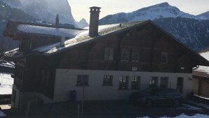 Un chalet traditionnel, doté d’un toit en pente, de nombreuses fenêtres et d’une cheminée, se dressant sur un fond de montagnes enneigées.