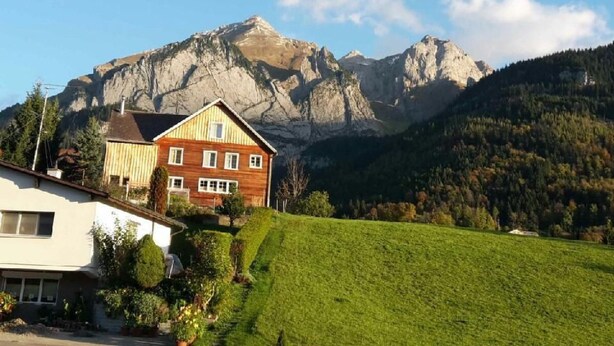 Ein zweistöckiges Holzhaus mit geneigtem Dach, auf einem Hügel mit Blick auf die Berge.