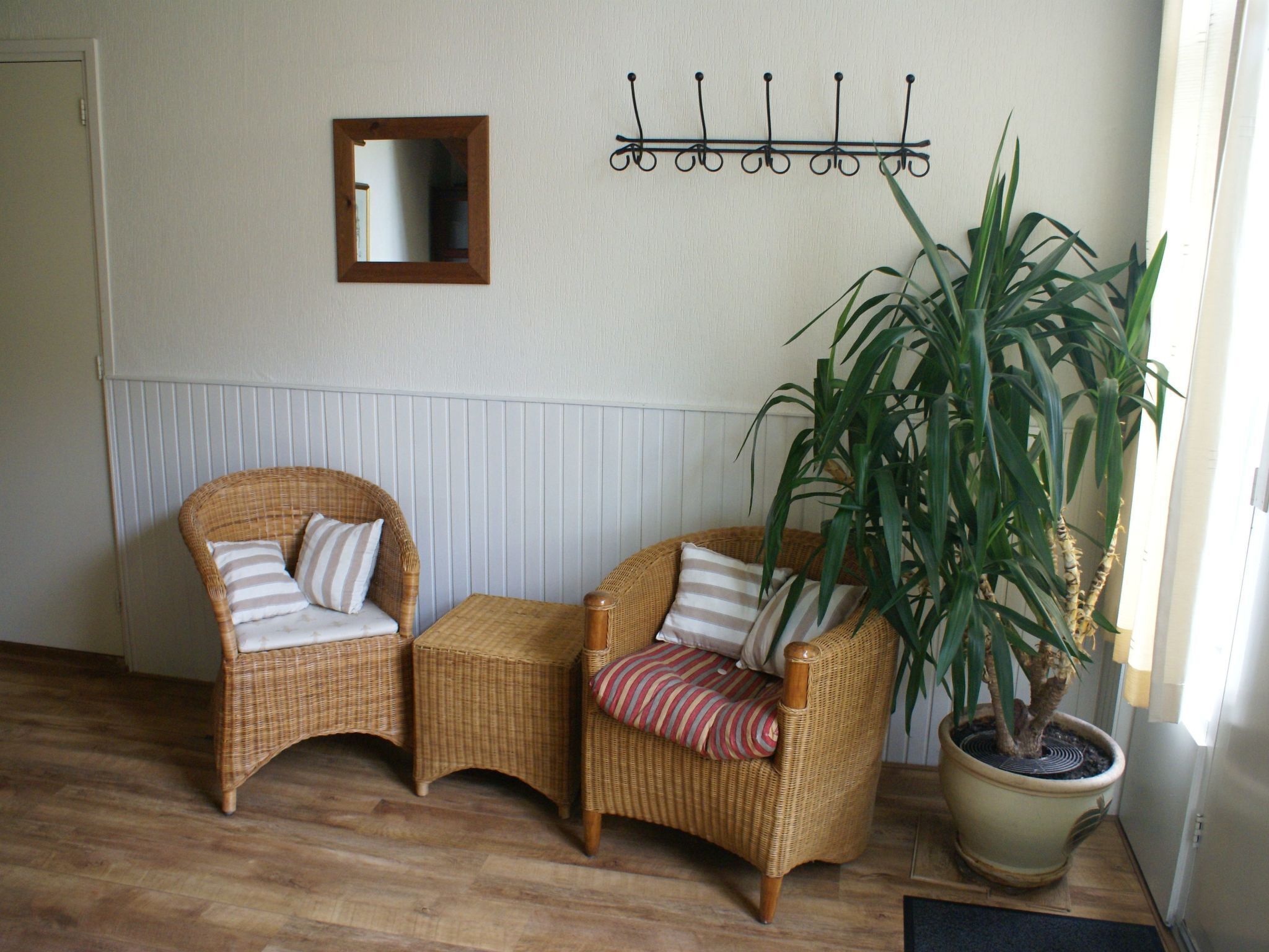 A room with two wicker chairs, a small table, a potted plant, a wall-mounted mirror, and a coat rack.