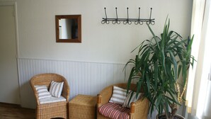 A room with two wicker chairs, a small table, a potted plant, a wall-mounted mirror, and a coat rack.